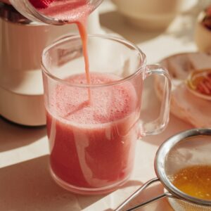 Delicious Strawberry Matcha Latte Recipe 15 Fresh strawberries being blended into a pink smoothie mixture, poured into a glass mug on a bright kitchen counter with honey and other ingredients nearby."
