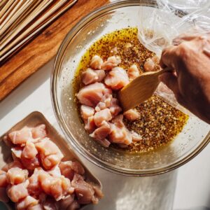 "A kitchen prep scene showing raw chicken pieces being mixed into a bowl of coconut and spice marinade beside a tray of bamboo skewers.