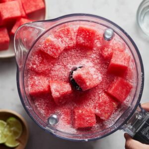 Best Watermelon Slushie Recipe 17 "Overhead view of a blender filled with bright red watermelon cubes mixed with crushed ice, with small bowls of water, sugar, and lime halves arranged around it on a light countertop."