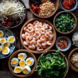 A top-down display of Pad Thai ingredients, including cooked shrimp, boiled eggs, bean sprouts, chopped herbs, chili flakes, peanuts, sauces, and fresh greens arranged in wooden and ceramic bowls.