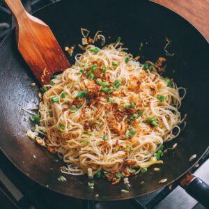 A wok filled with stir-fried rice noodles, topped with crispy shallots and chopped green onions, with a wooden spatula resting on the side.