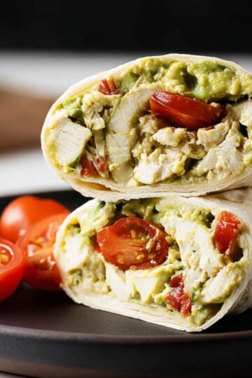 Healthy Avocado Chicken Wrap Recipe | 10-Minute Lunch 4 Avocado Chicken Wrap Close-up of an avocado chicken wrap cut in half on a dark plate, filled with shredded chicken, avocado, and cherry tomatoes.