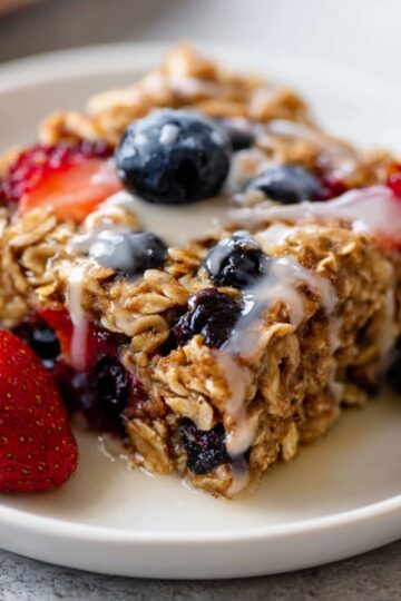 Easy Homemad Baked Oatmeal Recipe 22 Close-up of a slice of berry baked oatmeal drizzled with milk, topped with strawberries and blueberries on a white plate.