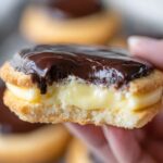 Boston Cream Pie Cookies Close-up of a Boston cream pie cookie held in hand, showing thick vanilla custard filling and glossy chocolate topping, soft background blur, warm bakery-style lighting”