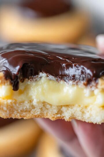 Boston Cream Pie Cookies Recipe | Easy & Delicious 1 Boston Cream Pie Cookies Close-up of a Boston cream pie cookie held in hand, showing thick vanilla custard filling and glossy chocolate topping, soft background blur, warm bakery-style lighting”