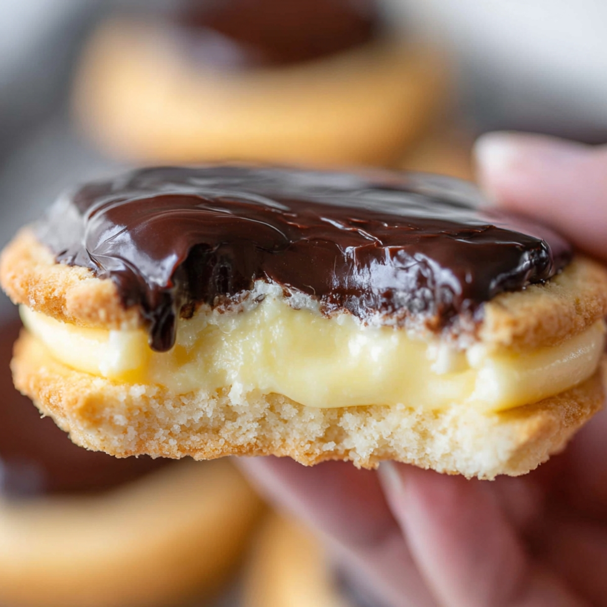 Boston Cream Pie Cookies Close-up of a Boston cream pie cookie held in hand, showing thick vanilla custard filling and glossy chocolate topping, soft background blur, warm bakery-style lighting”