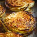 Golden Roasted Cabbage Steaks with charred edges and visible spice seasoning, showing layered cabbage texture in a swirl pattern. Macro food photography, dramatic warm lighting.