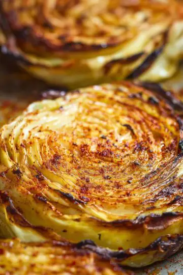 Easy Homemad Roasted Cabbage Steaks Recipe 23 Golden Roasted Cabbage Steaks with charred edges and visible spice seasoning, showing layered cabbage texture in a swirl pattern. Macro food photography, dramatic warm lighting.