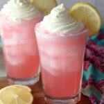 Whipped Pink Lemonade Two glasses of pink lemonade topped with whipped cream, garnished with lemon slices. The drinks are placed on a wooden board, and a colorful cloth is draped beside the glasses.