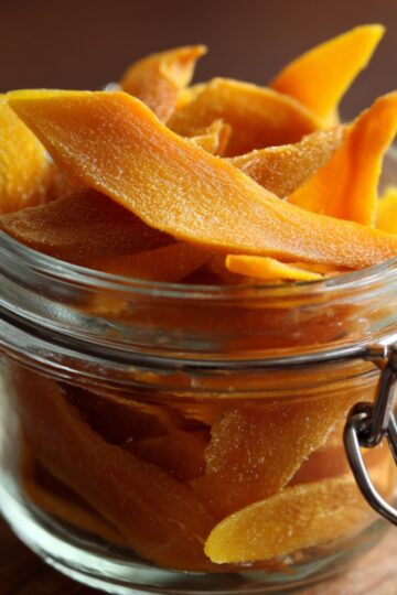 Dehydrated Mango A glass jar filled with bright orange dried mango slices, sitting on a wooden board with a metal clasp and a cooling rack in the background.