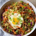 A bowl of Egg Roll In A Bowl featuring ground meat, shredded cabbage, carrots, and purple cabbage, topped with a crispy fried egg and green onions.