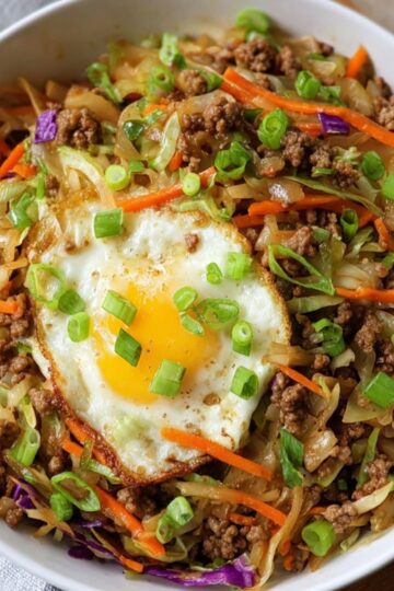 The Best Egg Roll In A Bowl Recipe 22 A bowl of Egg Roll In A Bowl featuring ground meat, shredded cabbage, carrots, and purple cabbage, topped with a crispy fried egg and green onions.