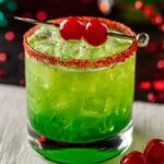 Grinch Cocktail Close-up of a vibrant green holiday cocktail filled with ice, topped with a red sugar rim and garnished with two red cherries on a cocktail pick, set against a colorful Christmas bokeh background.