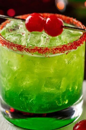 Grinch Cocktail Recipe - 5 Minutes, 6 Ingredients 18 Grinch Cocktail Close-up of a vibrant green holiday cocktail filled with ice, topped with a red sugar rim and garnished with two red cherries on a cocktail pick, set against a colorful Christmas bokeh background.