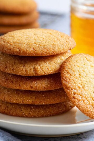 Easy Honey Cookies Recipe - Chewy, Spiced & Foolproof 8 Honey Cookies A stack of freshly baked golden honey cookies on a plate, with a honey jar and dipper in the background.