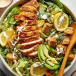 Honey Hot Chicken Salad Finished hot honey chicken Caesar salad with sliced crispy chicken, romaine lettuce, croutons, avocado slices, pumpkin seeds, lemon wedges, and creamy dressing in a large bowl.