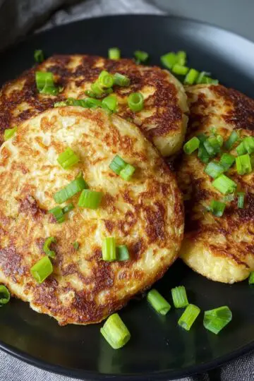 The Best Irish Potato Pancakes Recipe 7 Irish Potato Pancakes Three crispy golden boxty pancakes topped with green onions on a black plate. The pancakes are perfectly browned and fluffy inside.