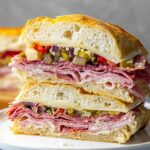 Italian Muffuletta Sandwich A thick slice of muffaletta sandwich stacked on a white plate, showing layers of salami, ham, provolone cheese, and olive salad.