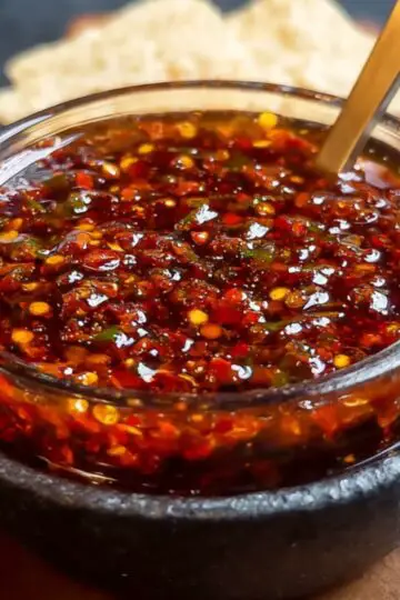A close-up of salsa macha in a small stoneware bowl, with chili flakes, oil, and spices visible. A golden spoon is placed inside, ready to stir the flavorful mixture.
