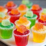 Sour Patch Jello Shots A close-up of colorful jello shots arranged in a group. The shots are topped with whipped cream and vibrant, gummy candy cubes in colors like orange, red, and green, making them visually appealing and fun for a party or gathering.