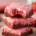 Strawberry Brownies Stacked strawberry brownies with pink glaze, one brownie bitten to show the moist and chewy texture inside.