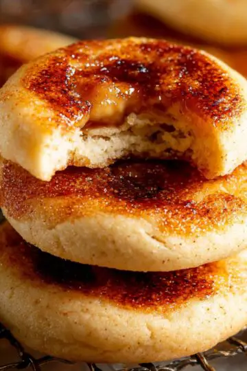 creme brûlée cookies stacked on a wire rack, topped with caramelized sugar, with one cookie bitten to show a soft buttery interior.