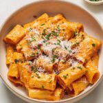 gochujang pasta Top-down view of creamy gochujang rigatoni pasta served in a beige bowl, garnished with grated cheese and green herbs on a light background.