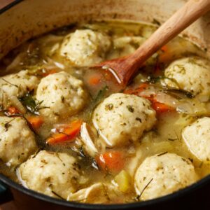 Easy Bisquick Chicken And Dumplings Recipe 17 A Dutch oven filled with chicken and dumplings simmering in broth, showing tender vegetables and fluffy Bisquick dumplings floating on top as a wooden spoon stirs the mixture.