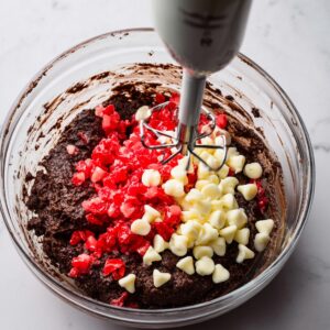 Black Forest Cookies Recipe | Easy Chocolate Cherry 16 A glass mixing bowl with cookie dough in the process of being mixed by an electric hand mixer. The dough contains chopped cherries and white chocolate chips