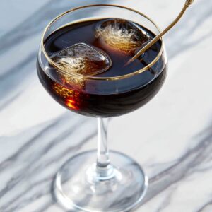 Delicious Black Manhattan Recipe 14 A Black Manhattan cocktail served in a gold-rimmed coupe glass with two clear ice cubes and a cocktail pick, sitting on a marble surface.