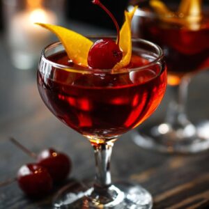 Delicious Black Manhattan Recipe 15 A Black Manhattan cocktail garnished with a Luxardo cherry and a twist of orange peel, served in a coupe glass in a dimly lit bar setting.
