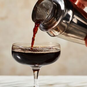 Delicious Black Manhattan Recipe 13 A bartender pouring a Black Manhattan cocktail from a metal shaker through a strainer into a chilled coupe glass on a marble countertop.