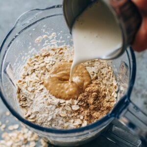 Milk being poured into a blender filled with rolled oats, nut butter, and dry ingredients for a blended oats recipe.