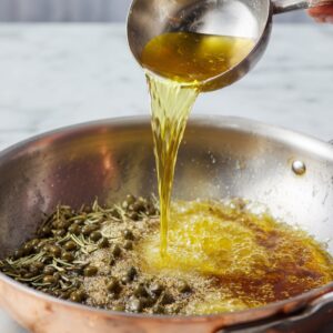 Healthy Chicken Piccata Recipe 16 A hand pours hot golden sauce from a metal ladle into a skillet filled with capers and herbs, creating a bubbling base for a lemon-caper pan sauce.