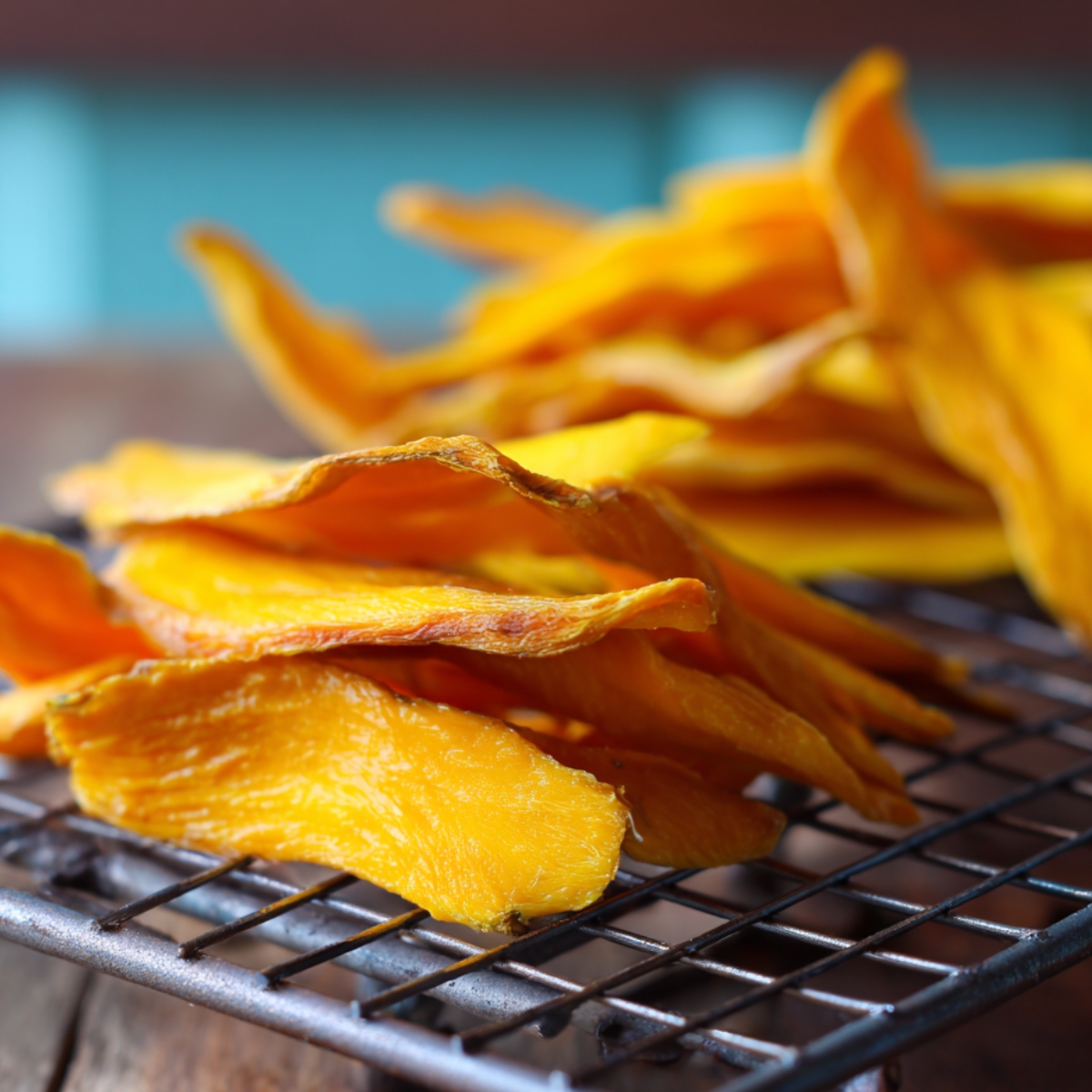 Thin dried mango slices spread out on a metal cooling rack, with more dried pieces piled behind on a rustic wooden surface.