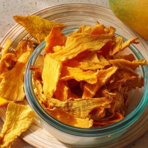 A glass jar filled with bright orange dried mango slices, sitting on a wooden board with a metal clasp and a cooling rack in the background.