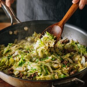 The Best Egg Roll In A Bowl Recipe 16 A large skillet filled with sautéed cabbage and ground meat, being stirred with a wooden spoon as the vegetables wilt and mix together.