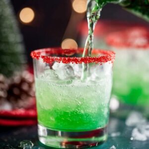 Grinch Cocktail Recipe - 5 Minutes, 6 Ingredients 14 Green festive cocktail being poured into an ice-filled glass with a red sugar rim, condensation on the glass, holiday lights softly blurred in the background for a cozy Christmas atmosphere.