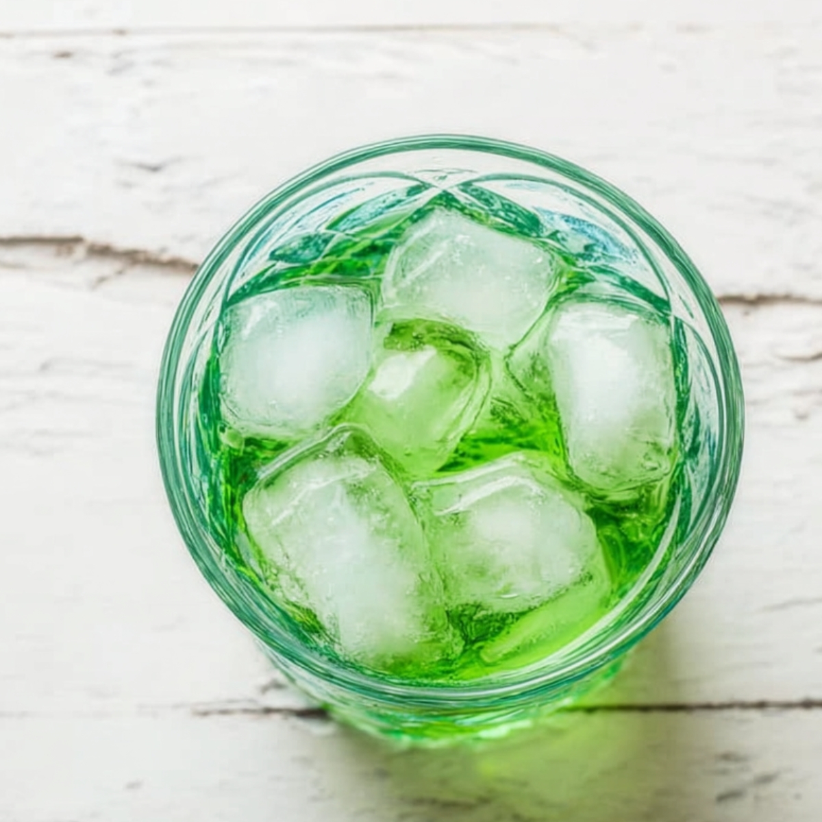 Grinch Cocktail Recipe - 5 Minutes, 6 Ingredients 10 Top-down view of a clear glass filled with ice cubes and bright green drink on a white wooden surface, minimalist food photography with clean lighting and simple composition.