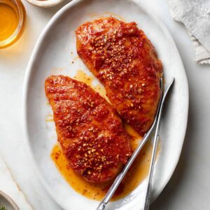 Honey Hot Chicken Salad Recipe | Crispy & Spicy in 30 Min 18 Two raw chicken breasts coated in spicy red marinade on a white oval plate, with metal tongs and small bowls of honey and spices nearby.