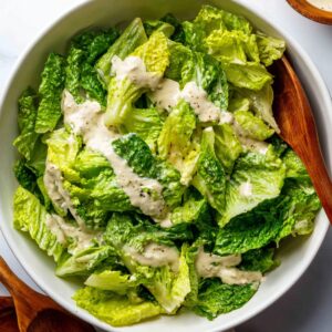 Honey Hot Chicken Salad Recipe | Crispy & Spicy in 30 Min 19 Chopped romaine lettuce tossed with creamy Caesar dressing in a white bowl, wooden salad spoons on the side.