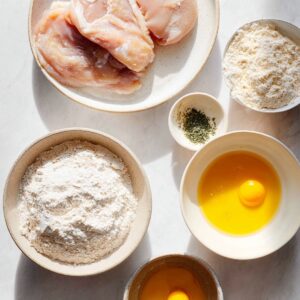 Honey Hot Chicken Salad Recipe | Crispy & Spicy in 30 Min 16 Overhead view of a breading setup with raw chicken breasts, bowls of flour, beaten egg, and dried herbs arranged on a light surface.
