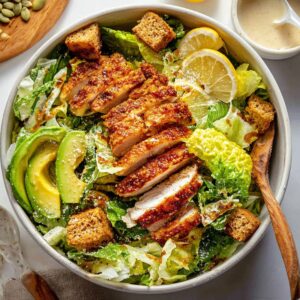 Honey Hot Chicken Salad Recipe | Crispy & Spicy in 30 Min 20 Finished hot honey chicken Caesar salad with sliced crispy chicken, romaine lettuce, croutons, avocado slices, pumpkin seeds, lemon wedges, and creamy dressing in a large bowl.