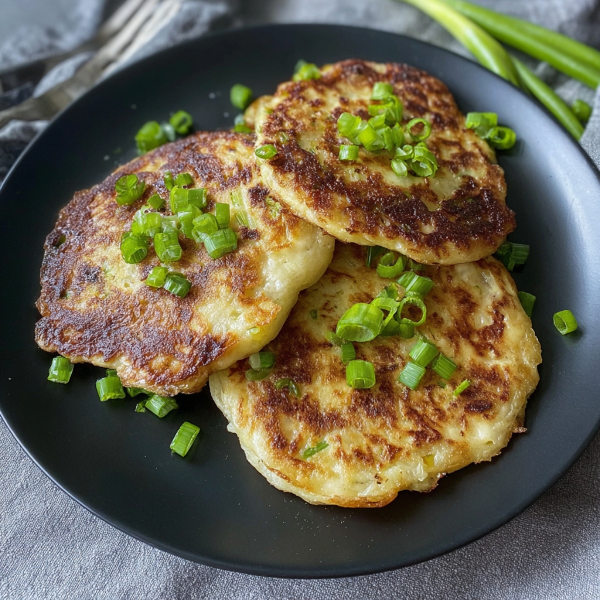 The Best Irish Potato Pancakes Recipe 14 Three crispy golden boxty pancakes topped with green onions on a black plate. The pancakes are perfectly browned and fluffy inside.