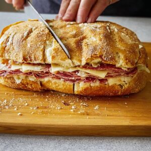 Easy Italian Muffuletta Sandwich | Ready in 15 Minutes 11 A large muffaletta sandwich on a wooden cutting board being sliced with a knife, showing layers of cured meats and cheese inside crusty bread.