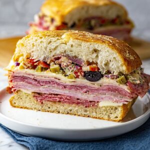 Easy Italian Muffuletta Sandwich | Ready in 15 Minutes 12 Italian Muffuletta Sandwich A thick slice of muffaletta sandwich stacked on a white plate, showing layers of salami, ham, provolone cheese, and olive salad.