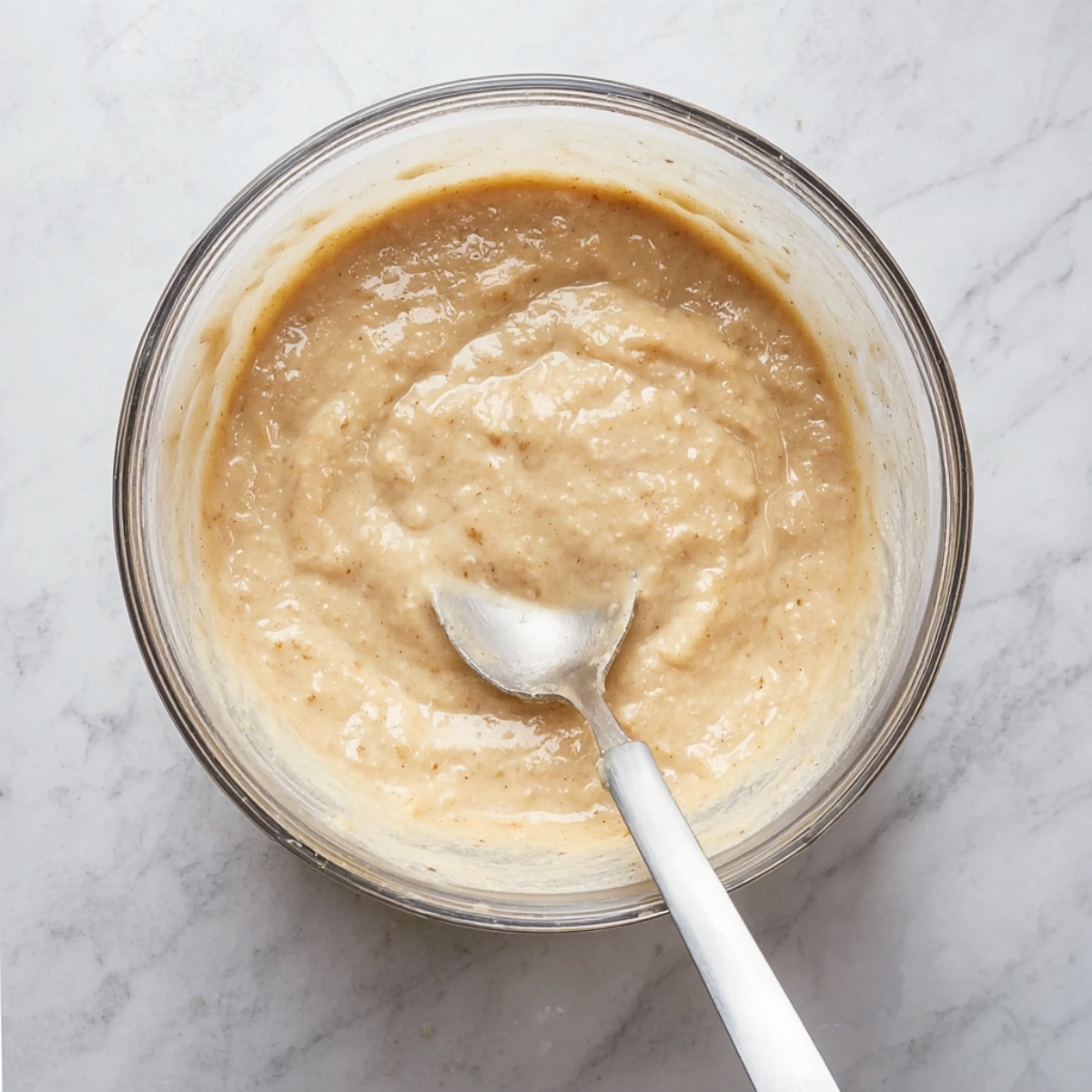 Easy Jamaican Banana Fritters Recipe | Ready in 15 Min 11 A close-up of banana fritter batter in a clear mixing bowl, showing a smooth and creamy texture with a spoon resting in it