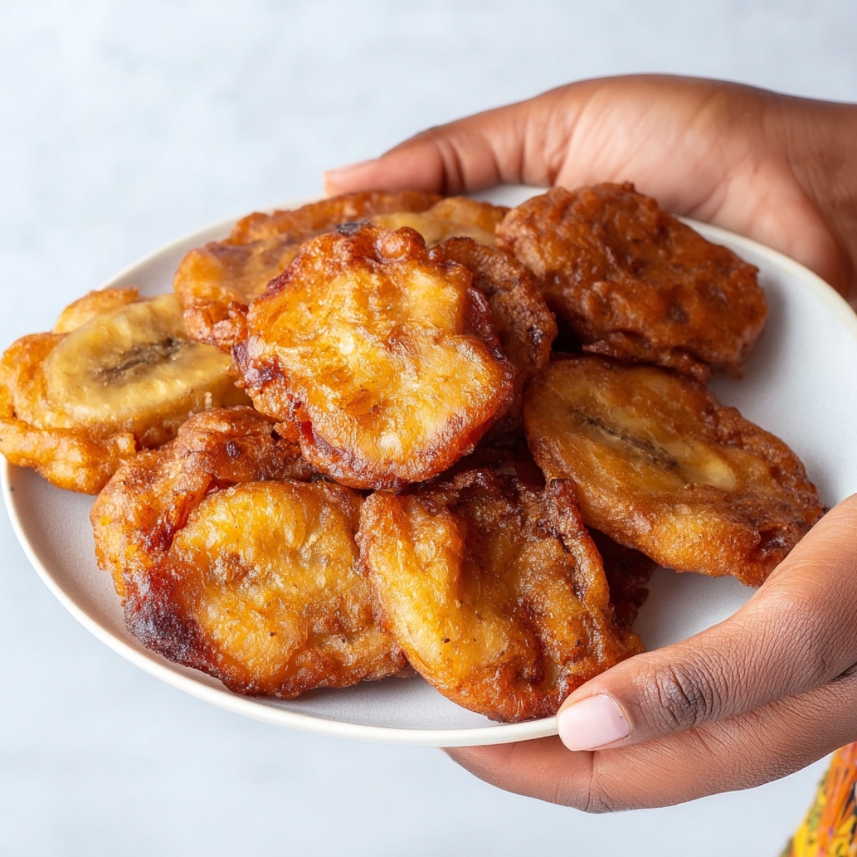 Easy Jamaican Banana Fritters Recipe | Ready in 15 Min 13 A hand holding a plate filled with golden brown Jamaican banana fritters. The fritters are crispy on the outside and soft on the inside, arranged neatly on the plate.