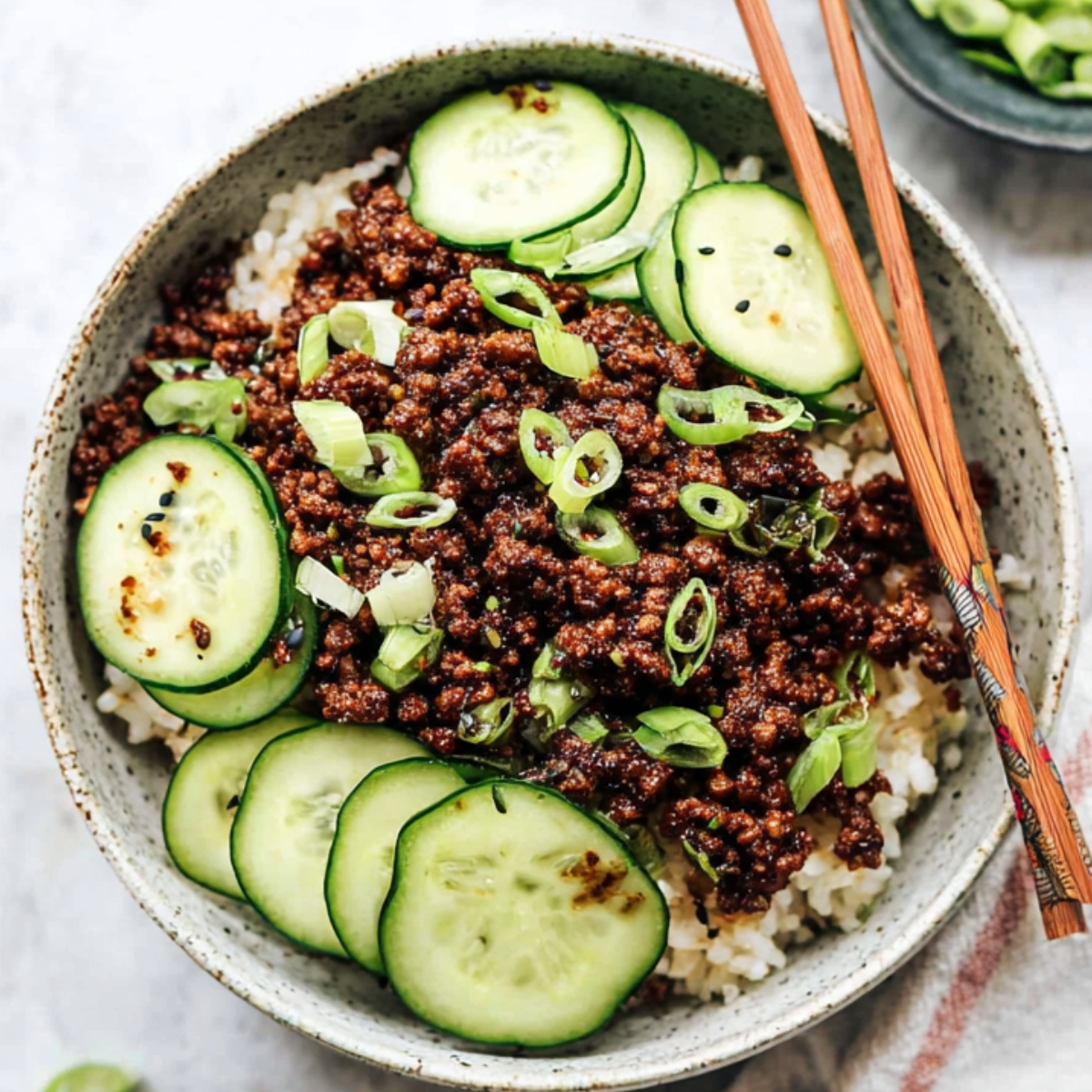 Easy Korean Ground Beef Bowl Recipe | 20-Minute Dinner 13 Korean ground beef served over white rice, topped with sliced cucumbers and green onions, with chopsticks on the bowl.