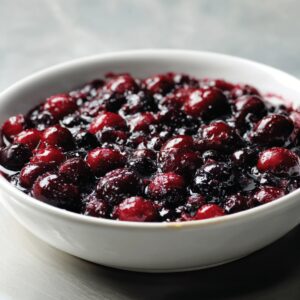 Close-up of a white bowl filled with mixed berries coated in sugar and juice, creating a glossy, vibrant berry filling.